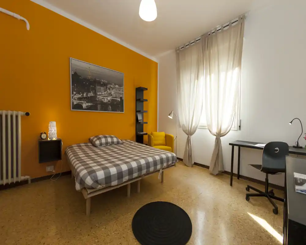 Bright, well-staged bedroom with an orange accent wall, double bed with checked bedding, a comfy armchair and a desk by the window — ideal for a cozy single or couple.