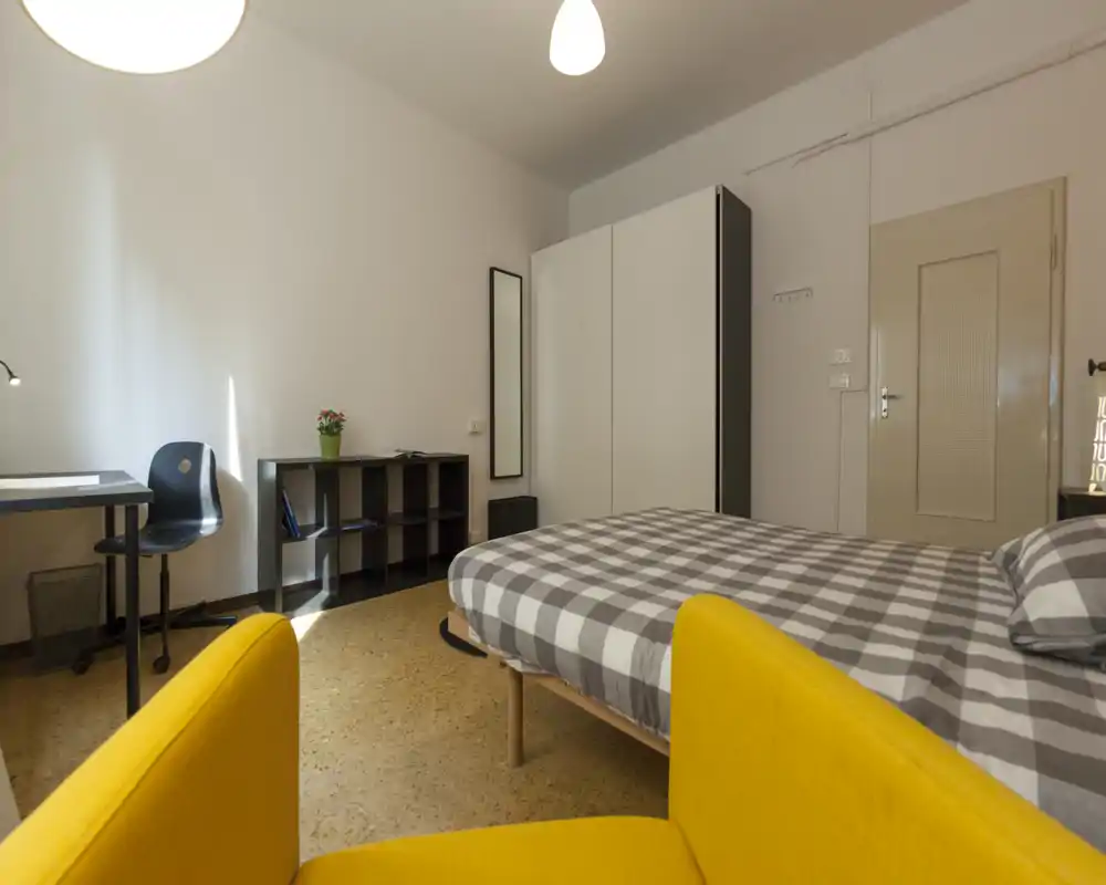 Bright, well-composed bedroom with a double bed in checked linen, a yellow armchair in the foreground and a compact desk and wardrobe along the wall.
