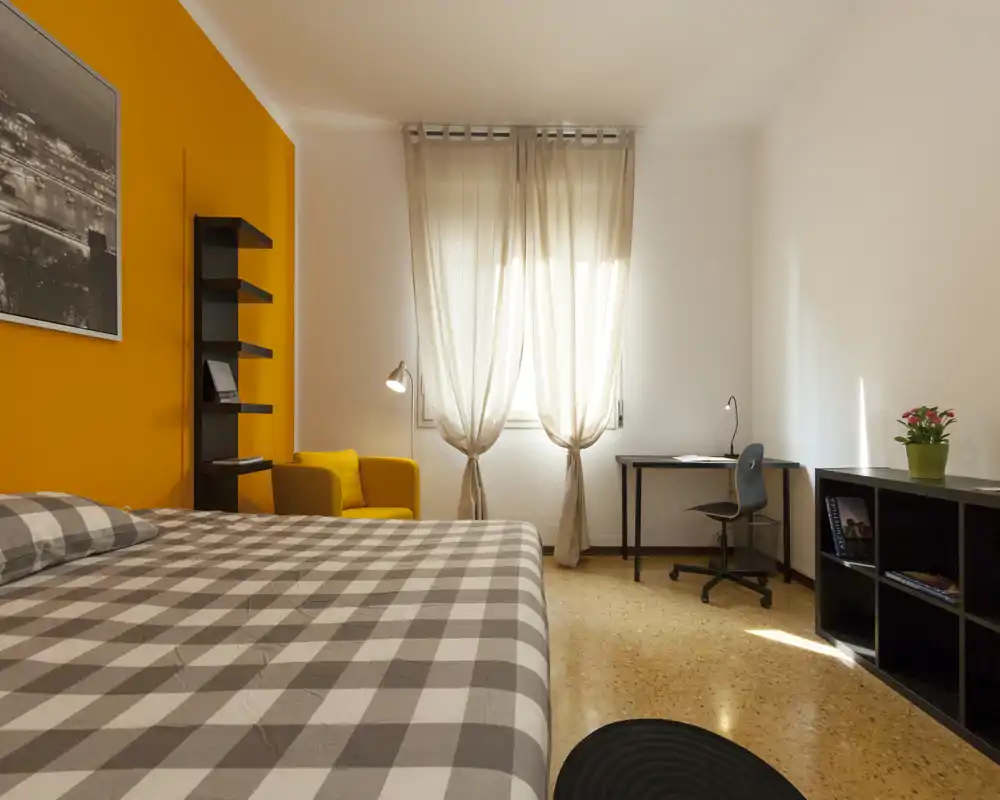 Bright, well-composed bedroom with a large checked bed in the foreground, a cozy yellow accent wall and armchair, a desk by the window and shelving—ideal for photos for a rental listing.