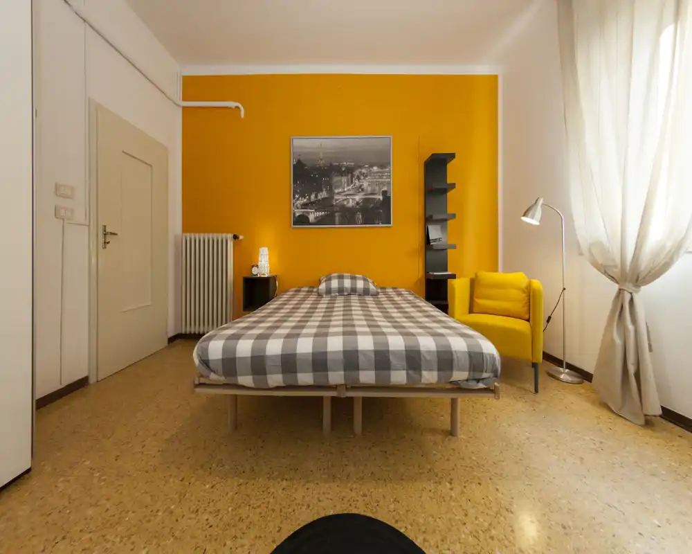 Bright, well-staged bedroom with a central double bed against a warm orange accent wall, a cozy yellow armchair and natural light from a large window.