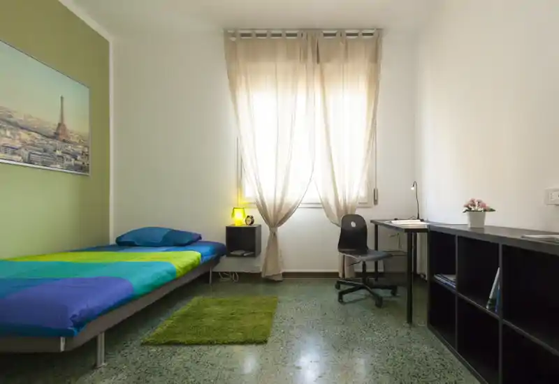 Dormitorio individual luminoso y ordenado con cama baja, lámpara de noche y un pequeño escritorio junto a la ventana — ideal para estudiante o una sola persona.