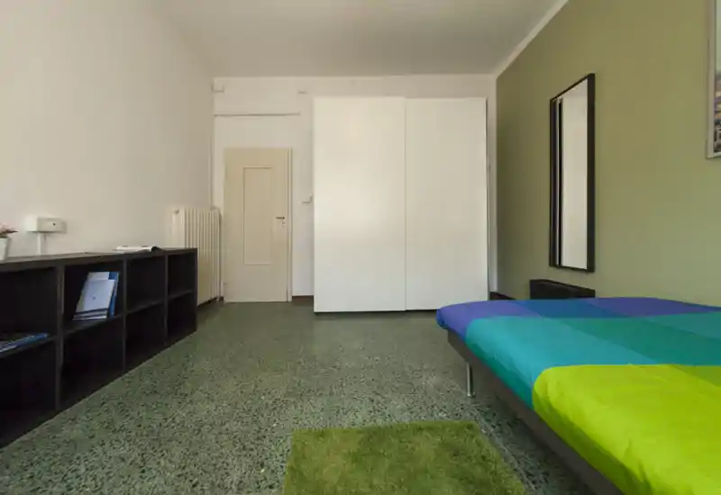 Dormitorio sencillo y luminoso con una cama individual de colores, espejo en la pared y un gran armario blanco. Estanterías minimalistas y una alfombra verde completan el espacio ordenado.