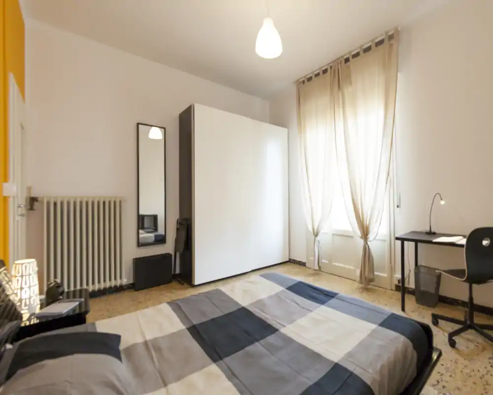 Camera da letto luminosa e curata con letto matrimoniale, grande armadio, scrivania e luce naturale soffusa dalla finestra con tende.