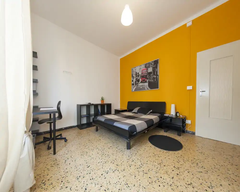 Camera da letto luminosa e moderna con letto matrimoniale contro una parete arancione, comodini minimal e una piccola scrivania vicino alla finestra. Pavimento in terrazzo lucidato e quadro decorativo donano un aspetto fresco e accogliente.