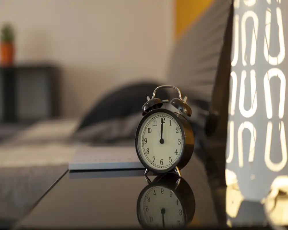 Primo piano del comodino in una camera da letto con una sveglia classica, la biancheria morbida sullo sfondo e una luce calda che crea un'atmosfera accogliente.