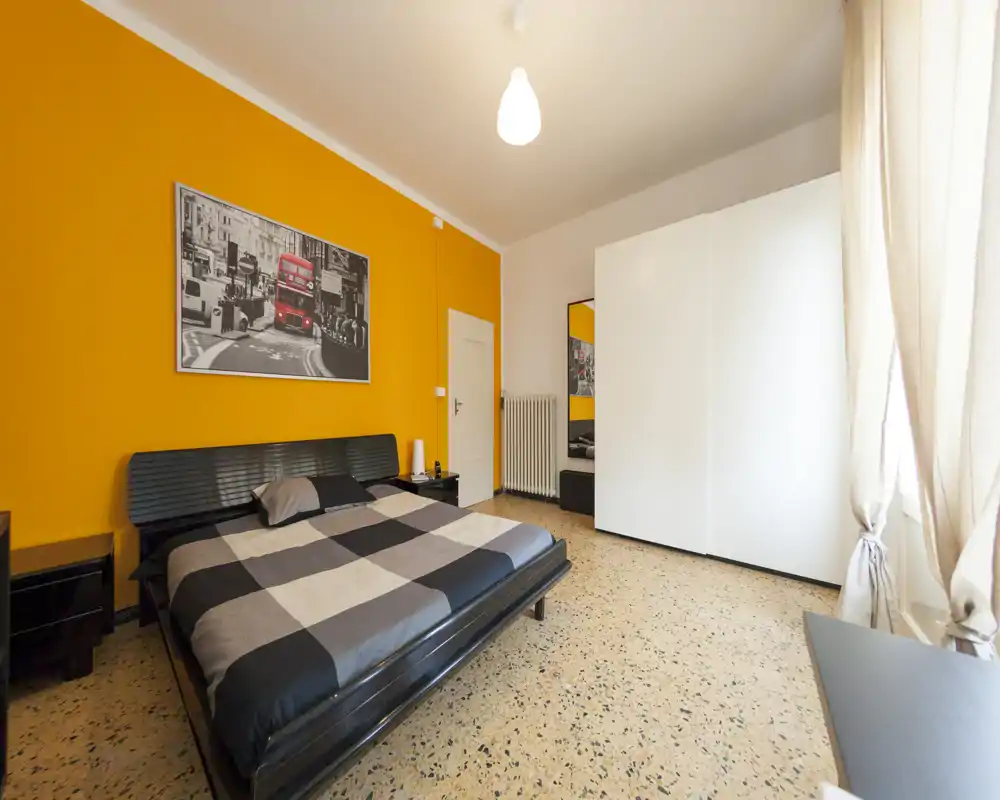 Camera da letto luminosa e moderna con un grande letto matrimoniale, parete accentuata arancione e un ampio armadio bianco. La luce naturale filtra dalle tende leggere, creando un ambiente caldo e accogliente, perfetto per annunci di affitto.