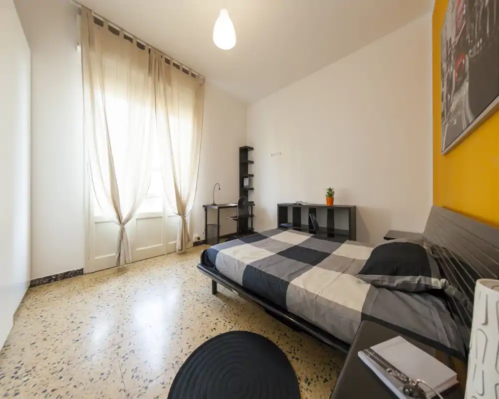 Camera da letto luminosa e ordinata con letto matrimoniale, scrivania e mensole. Ampia finestra con tende leggere che garantisce molta luce naturale e un aspetto moderno.