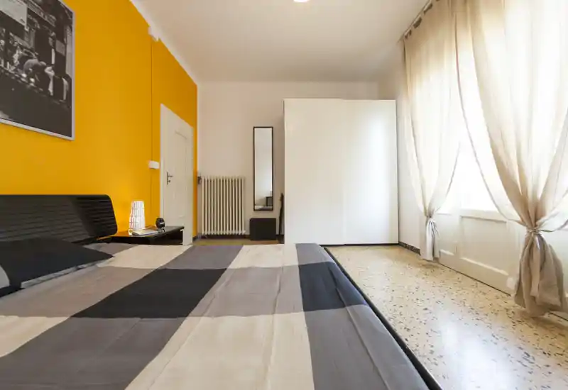 Camera da letto luminosa e moderna con un ampio letto in primo piano, parete accentata gialla, armadio e tende leggere che filtrano la luce naturale.