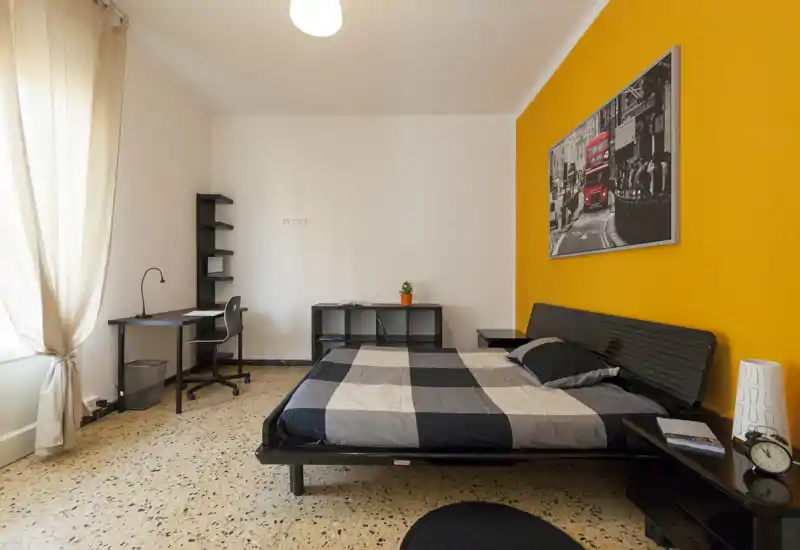 Camera da letto luminosa e ben arredata con letto matrimoniale, scrivania e parete d'accento gialla. Ampia finestra per luce naturale, comodini e un quadro decorativo.