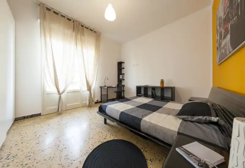 Camera da letto luminosa e ordinata con letto matrimoniale, scrivania e mensole. Ampia finestra con tende leggere che garantisce molta luce naturale e un aspetto moderno.
