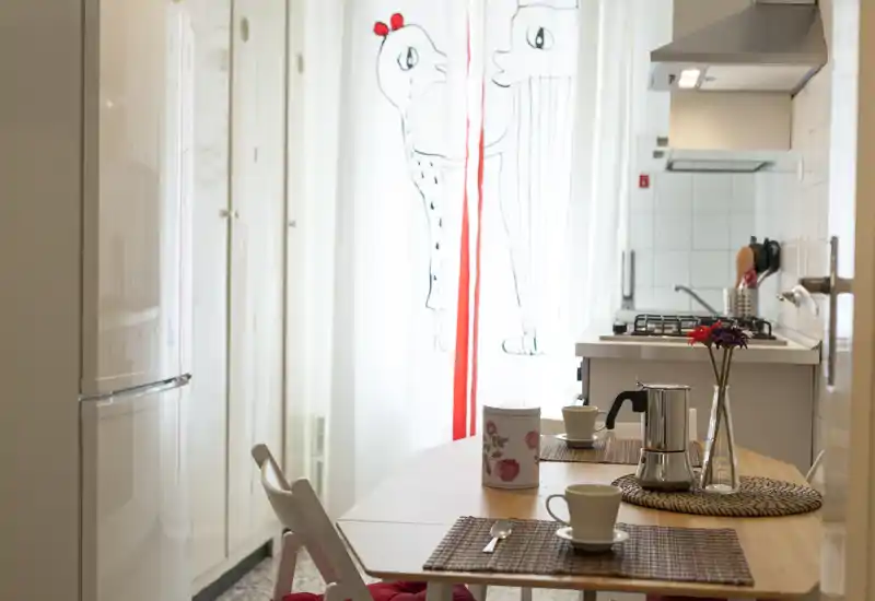 Cucina compatta e luminosa con tavolo da pranzo per il caffè, sottopiatti decorativi e piano cottura a gas moderno; luce naturale morbida dalle tende illustrate.