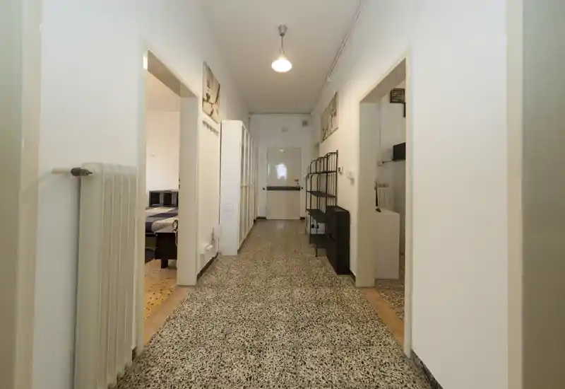 Lungo corridoio/ingresso ben illuminato con pavimento in graniglia, pareti bianche e più porte che si affacciano su stanze adiacenti; mensole e termosifone lungo le pareti.