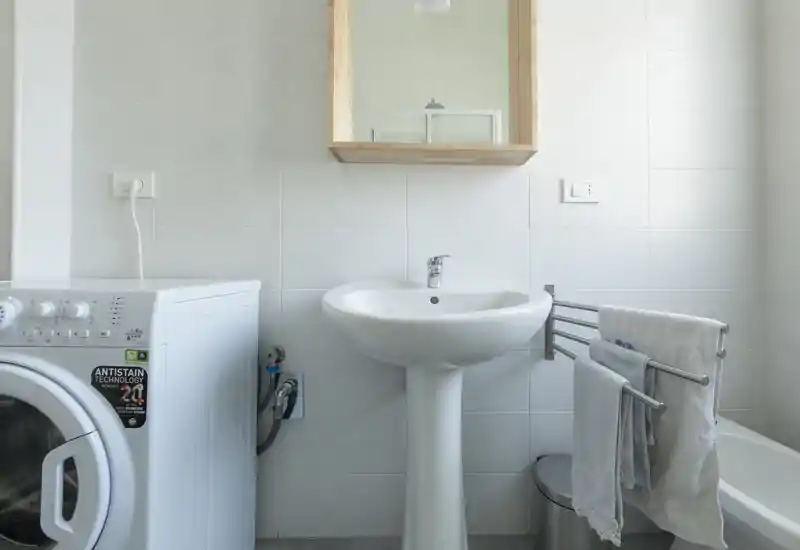 Bagno luminoso e pulito con lavabo a colonna, specchiera e lavatrice a sinistra; piastrelle bianche e porta asciugamani offrono un ambiente funzionale e ordinato.