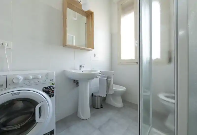Bagno compatto e luminoso con lavabo a colonna, wc, doccia con vetro e lavatrice; piastrelle neutre e luce naturale dalla finestra.