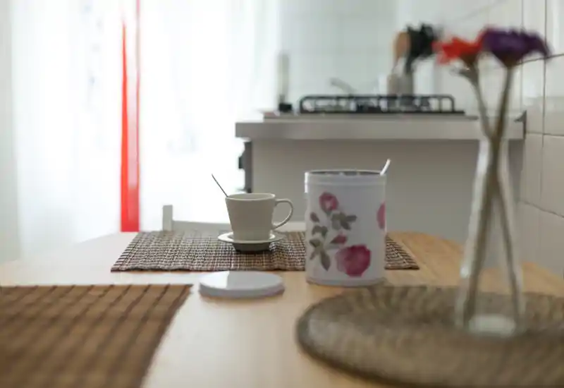 Angolo cucina luminoso e ben illuminato con tavolo in legno apparecchiato con una tazza, una scatola decorativa e sottopiatti; un piano cottura a gas è visibile sullo sfondo.