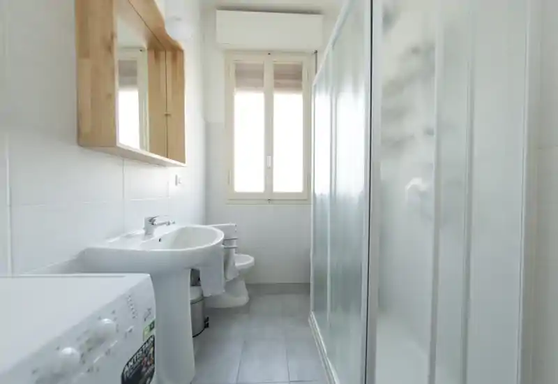 Bagno pulito e luminoso con lavabo a colonna, bidet, box doccia in vetro satinato e luce naturale dalla finestra.
