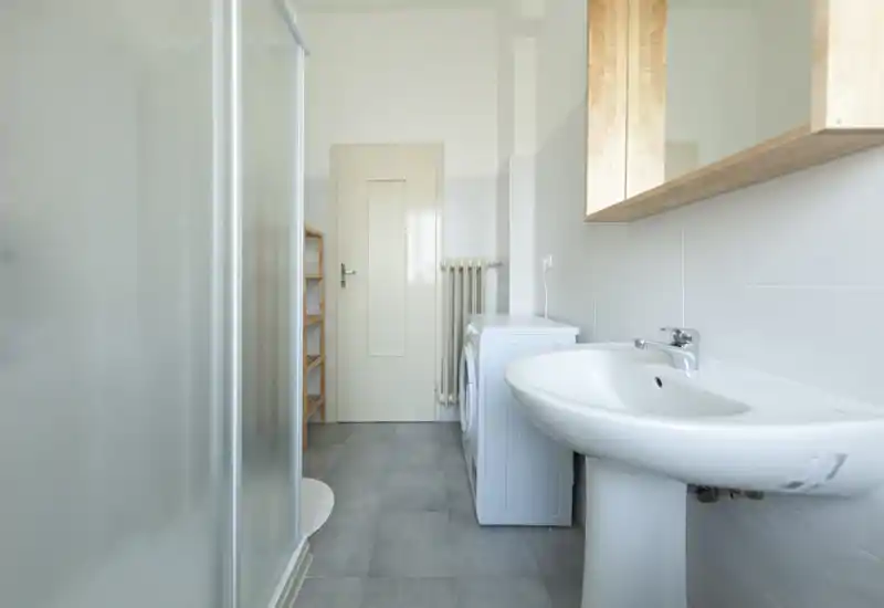 Bagno luminoso e moderno con lavabo a colonna, cabina doccia e lavatrice — piastrelle neutre e linee pulite per un ambiente funzionale.