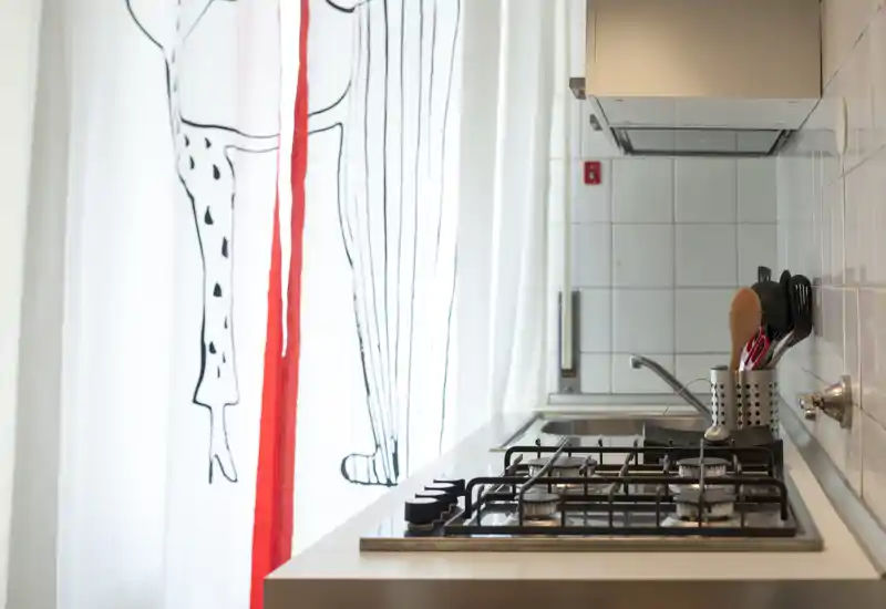Angolo cucina moderno e compatto con piano cottura a gas, lavello in acciaio e portautensili, illuminato da una tenda decorata.