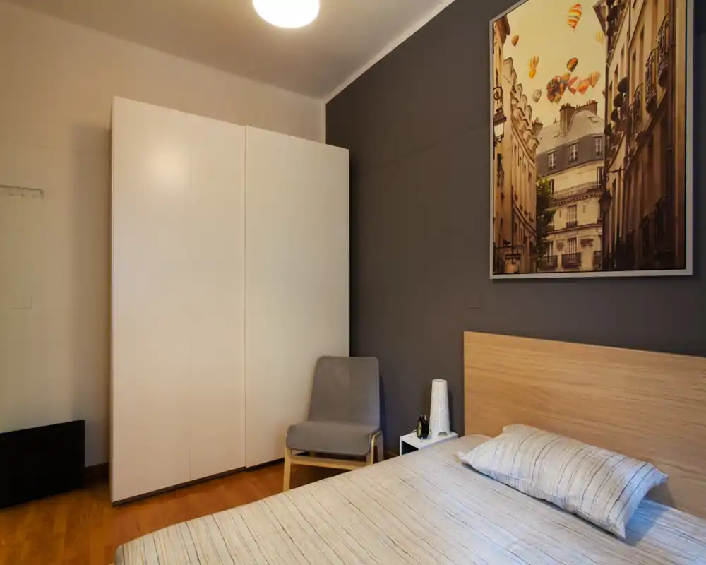 Bright, neatly styled bedroom with a double bed, wooden headboard, wardrobe and chair; neutral linens and framed wall art create a calm, modern atmosphere.