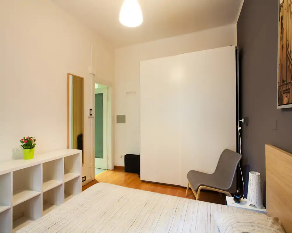 Sunny, well-styled bedroom with a large bed, white wardrobe and a comfortable lounge chair; warm wooden floors and soft lighting create a welcoming atmosphere.
