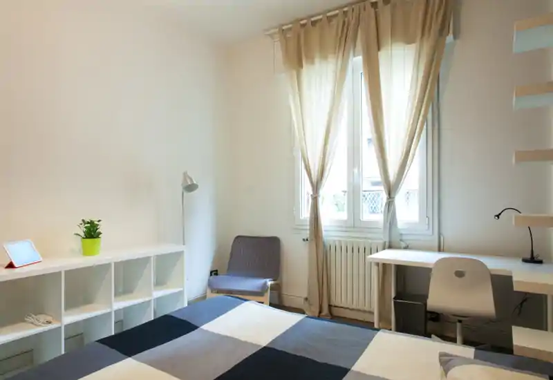 Bright, minimalist bedroom with a large bed, desk by the window and cozy accents like a lounge chair and curtains.
