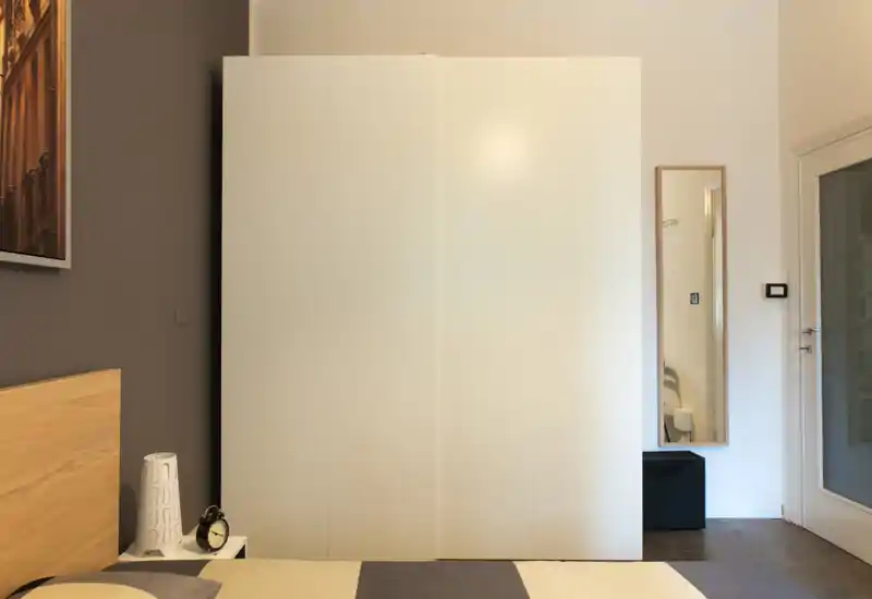 Calm, minimal bedroom view with a wooden headboard, bedside lamp and alarm clock; a large white wardrobe and a full-length mirror create a neat, modern feel.