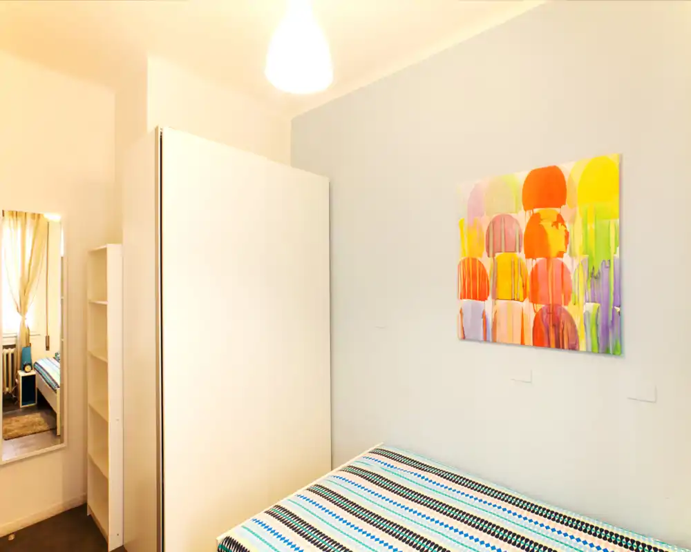 Camera da letto compatta e luminosa con copriletto a righe, armadio bianco e quadro colorato; arredamento minimal e illuminazione calda.
