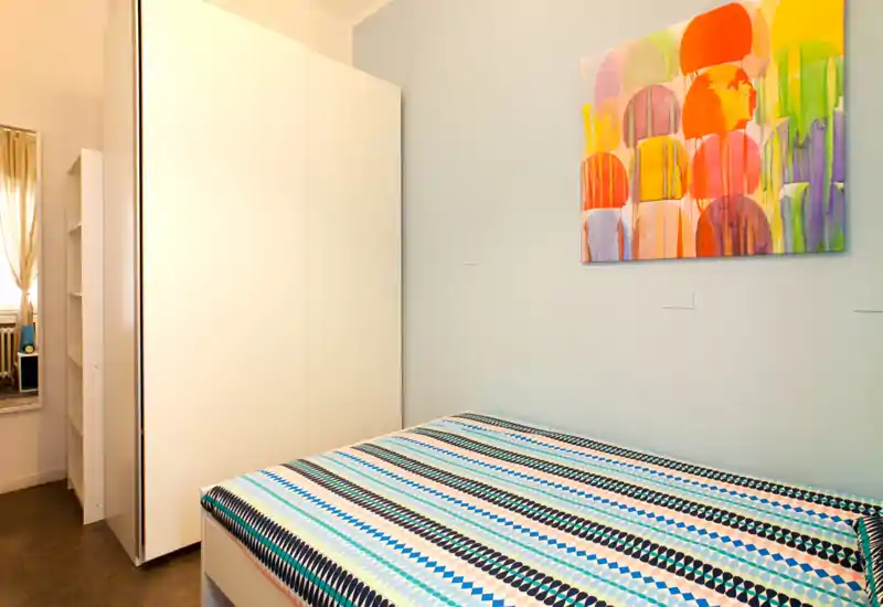 Camera da letto compatta e luminosa con letto matrimoniale a fantasia, grande armadio bianco e quadro colorato che dona un aspetto moderno.