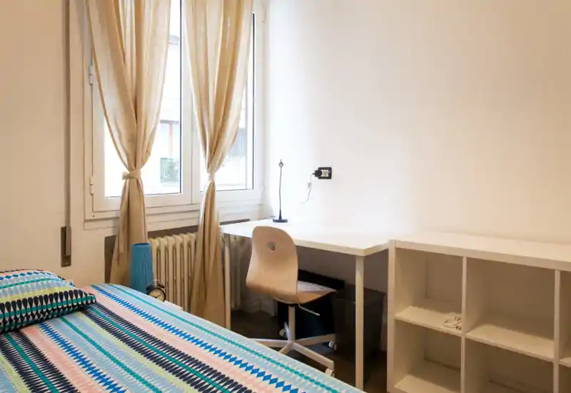 Camera da letto luminosa e ordinata con copriletto fantasia, scrivania e scaffalatura vicino alla finestra che offre luce naturale — ideale per lavoro e riposo.