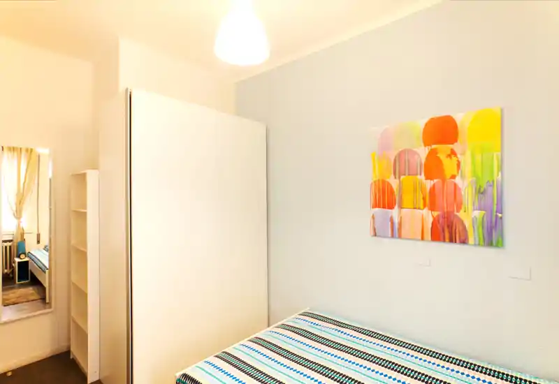 Camera da letto compatta e luminosa con copriletto a righe, armadio bianco e quadro colorato; arredamento minimal e illuminazione calda.
