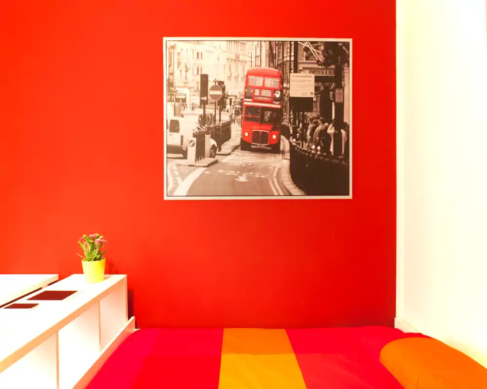 Camera da letto vivace con letto colorato e parete rossa decorativa con quadro urbano; ambiente luminoso, accogliente e di stile, perfetto per gli annunci.