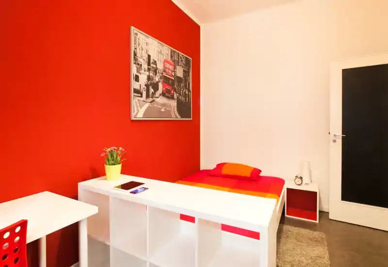 Camera singola moderna e luminosa con parete rossa intensa, letto ordinato con biancheria colorata, scrivania e scaffalatura — ideale per studenti o ospiti singoli.