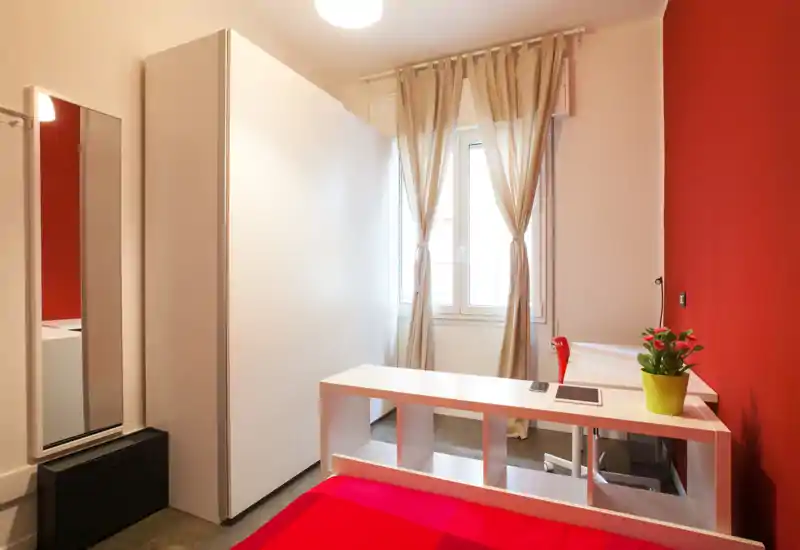 Camera da letto compatta e ben arredata con parete d'accento rossa, mobilio bianco e finestra che illumina naturalmente. Il letto è rifatto, c'è una scrivania/divisorio e una pianta decorativa.