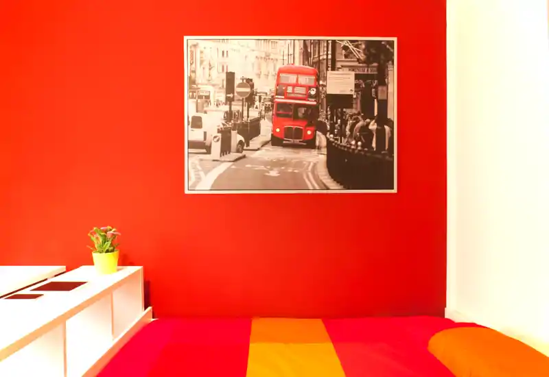 Camera da letto vivace con letto colorato e parete rossa decorativa con quadro urbano; ambiente luminoso, accogliente e di stile, perfetto per gli annunci.