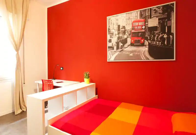 Camera da letto moderna e luminosa con copriletto colorato, divisorio contenitore bianco e una piccola scrivania vicino alla finestra; parete accent rossa e stampa cittadina.