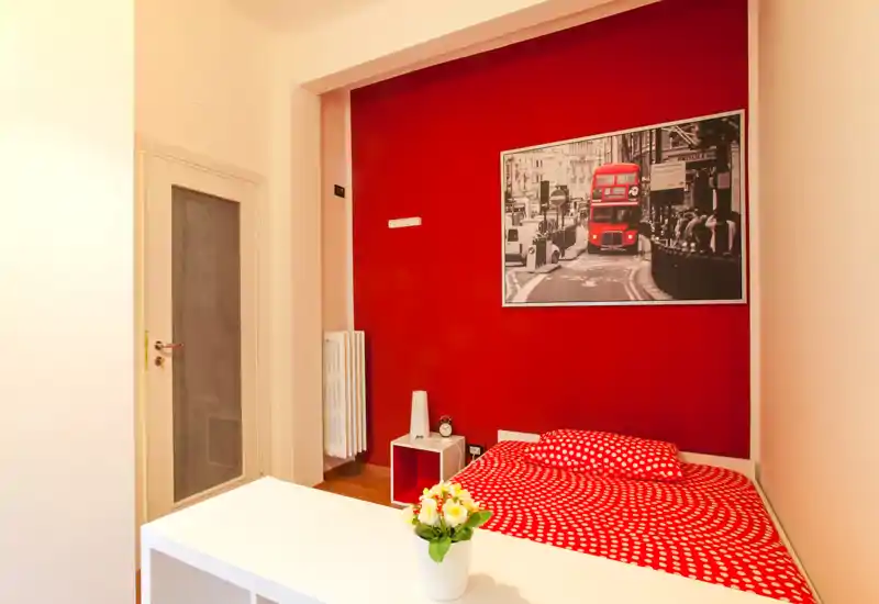 Camera singola luminosa e di design con parete rossa a contrasto e biancheria coordinata a pois. Include un piccolo comodino, una stampa decorativa e una scrivania minimal con fiori, ideale per un soggiorno urbano accogliente.