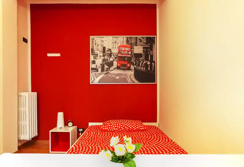 Camera compatta con parete accentata rosso vivo e copriletto a pois, illuminazione luminosa e comodino minimal con elementi decorativi.