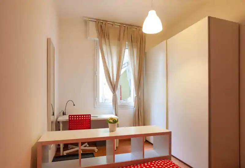 Camera da letto compatta con piccolo letto parzialmente visibile, scrivania bianca vicino alla finestra, ampia armadiatura e arredi moderni semplici. Spazio luminoso e ordinato con toni neutri e accenti rossi.