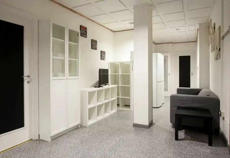 Piccola zona living con divano grigio, mensole modulari bianche e una TV — stile pulito e neutro con illuminazione uniforme, adatto a un monolocale.