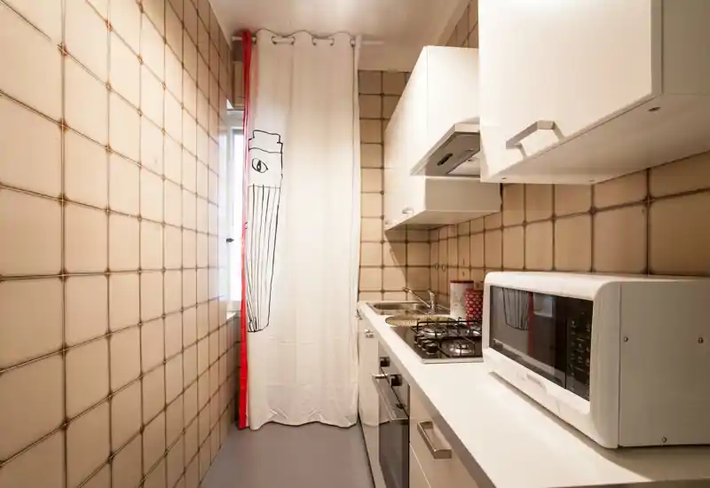 Compact galley kitchen with white cabinets, gas hob, microwave and a decorative curtain at the window; bright and tidy space ideal for short-stay listings.