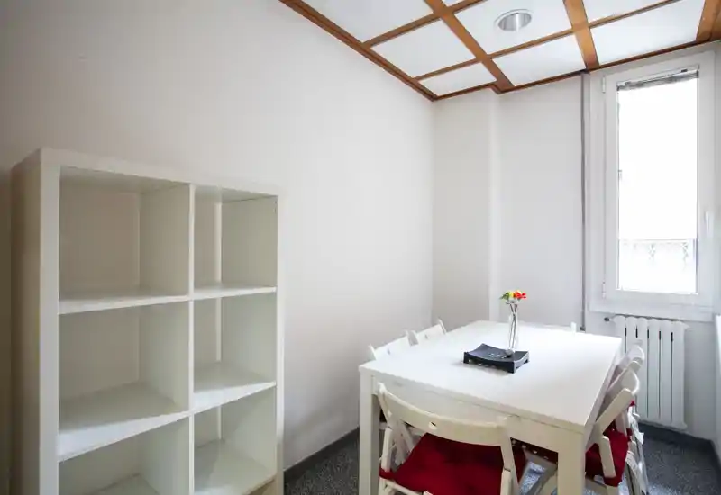 Compact, bright dining area with a white table, four chairs with red cushions and a decorative vase; clean minimalist shelving and natural light from a window.