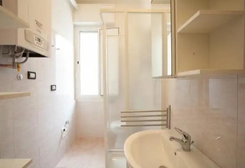 Clean, bright bathroom with a sink, modern faucet, enclosed shower and wall-mounted shelving — neutral tiled walls and a window for natural light.