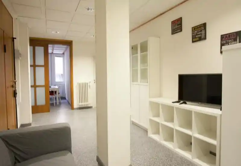 Tidy living area with a compact sofa, TV on a low white shelving unit and a clean, bright layout leading to a dining nook. Neutral tones and minimal decoration create a simple, functional living room ideal for short-term rentals.