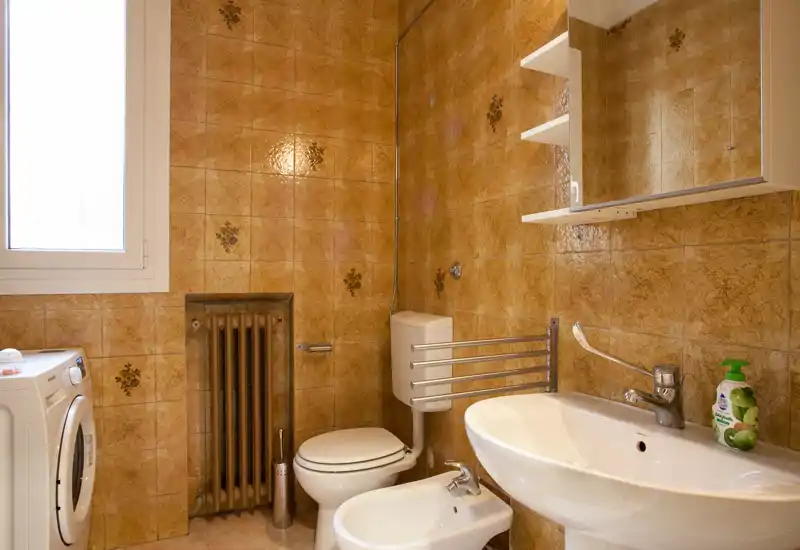Clean, well-lit bathroom with sink, bidet, toilet and shower area; warm patterned wall tiles and a mirrored cabinet provide storage and reflect light.