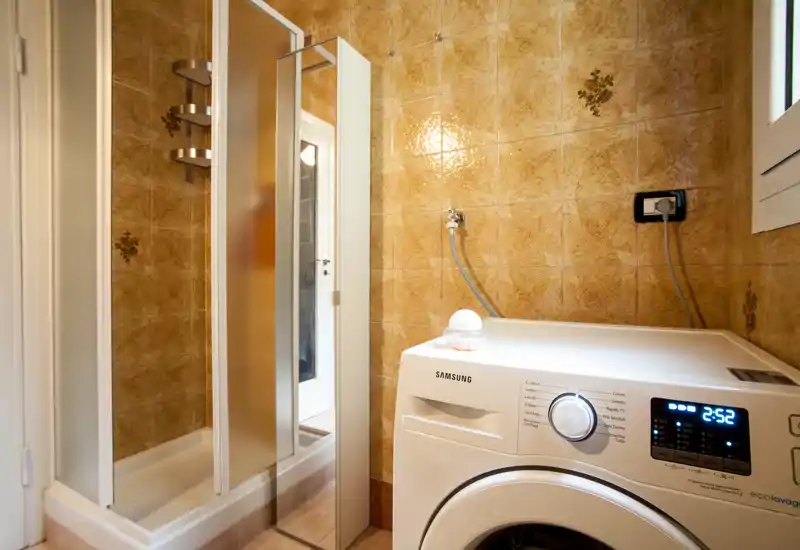 Compact bathroom with a corner shower and a front-loading Samsung washing machine; warm tiled walls and a small window provide natural light.