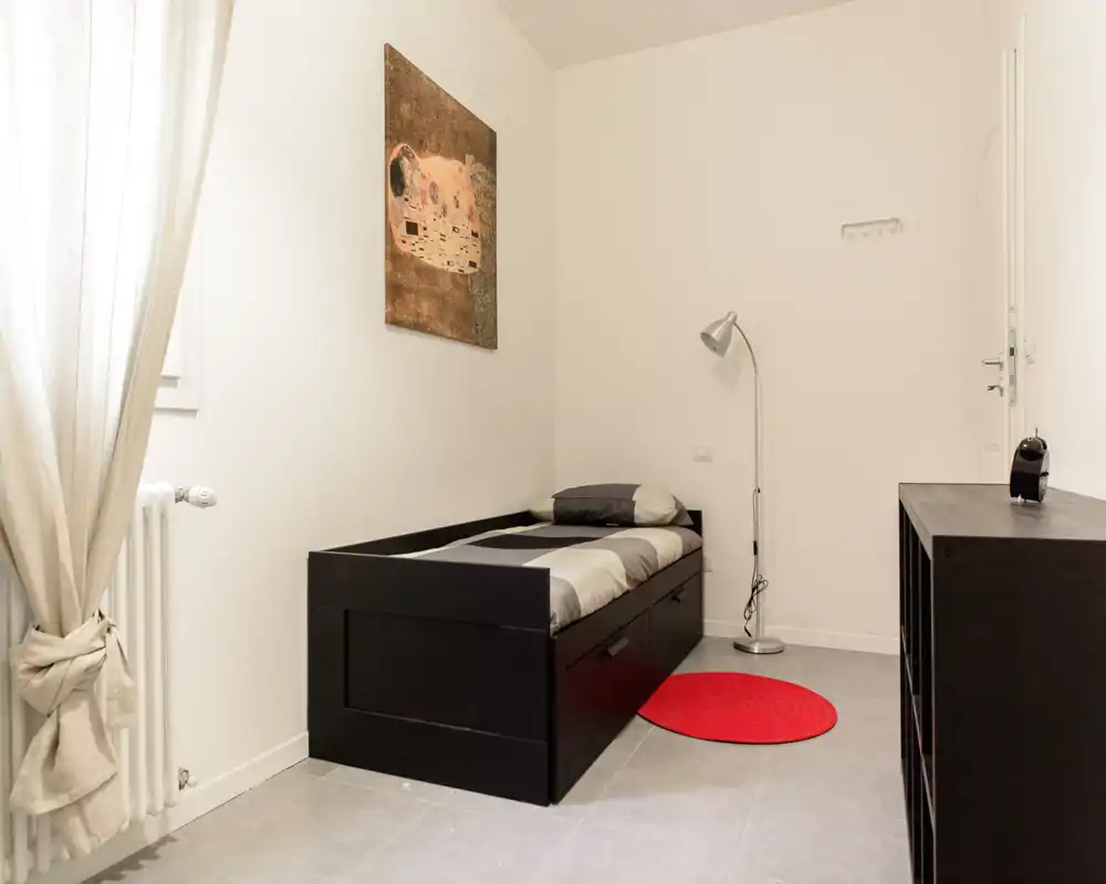 Camera singola compatta con letto nero, lampada da terra e arredo minimal; ambiente luminoso e pulito con toni neutri e un tappeto rosso come accento.