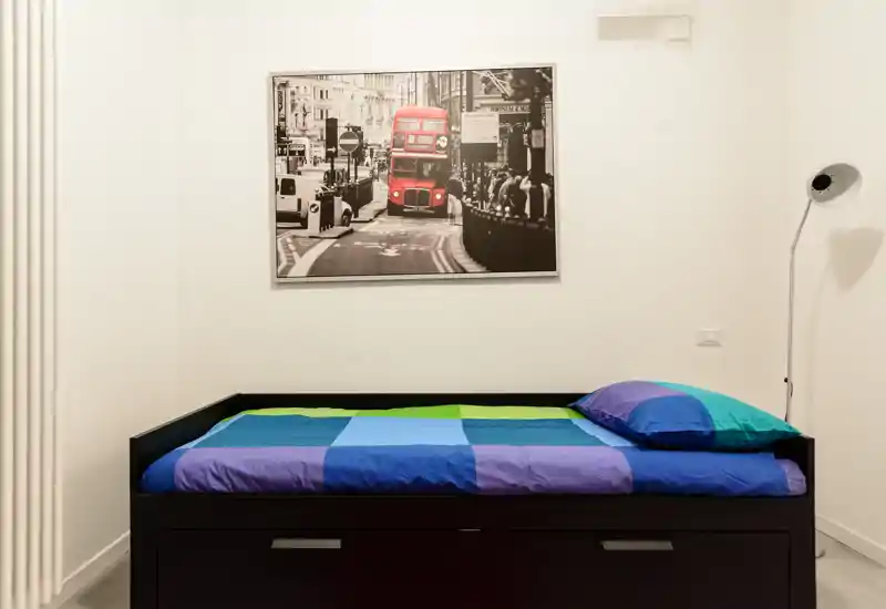Camera singola compatta con letto ben rifatto, biancheria colorata e una stampa incorniciata sopra. Spazio ordinato e ben illuminato, ideale per un ospite singolo o uno studente.