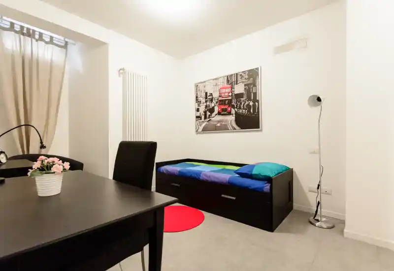 Piccola camera arredata con un daybed singolo con cassetti contenitore, biancheria colorata, lampada da terra e quadro alle pareti. Stile pulito e moderno con scrivania in primo piano e pareti neutre che rendono lo spazio luminoso e funzionale.
