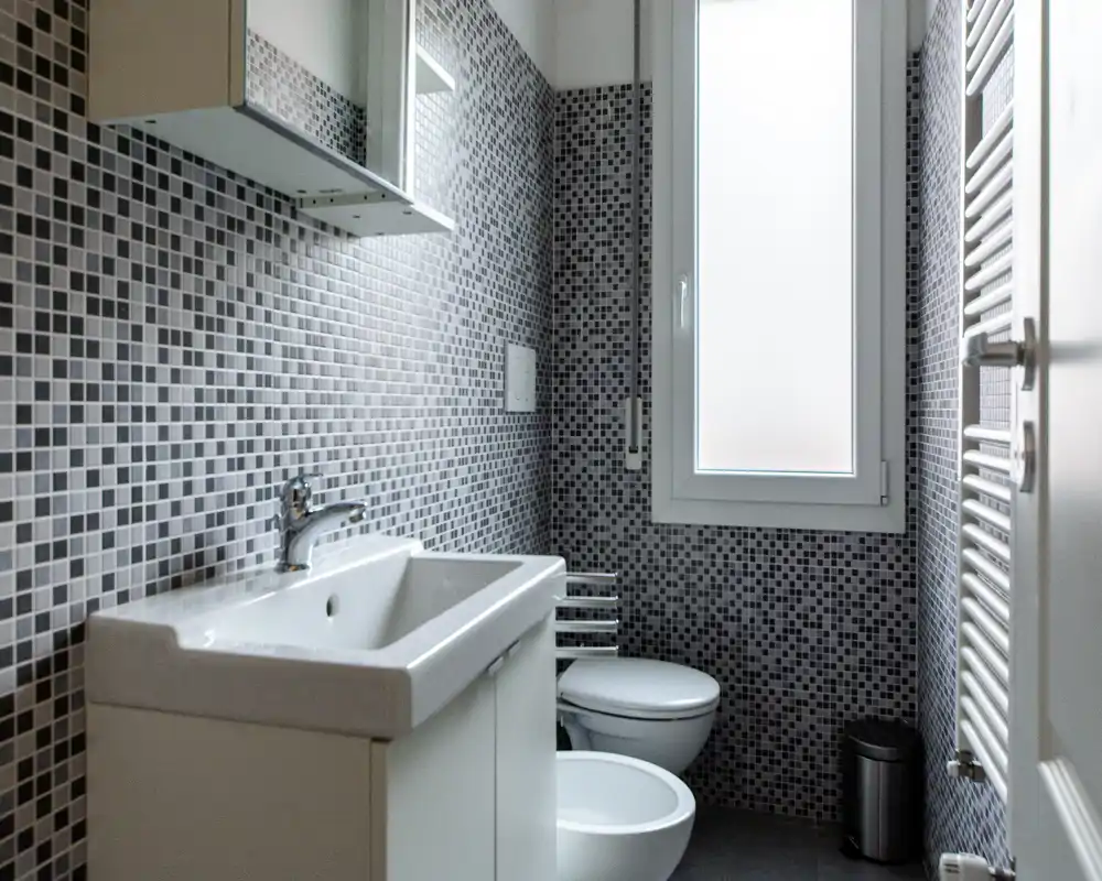 Bagno moderno e compatto con pareti in piastrelle a mosaico, mobile lavabo, bidet e WC, e una finestra satinata che illumina naturalmente.