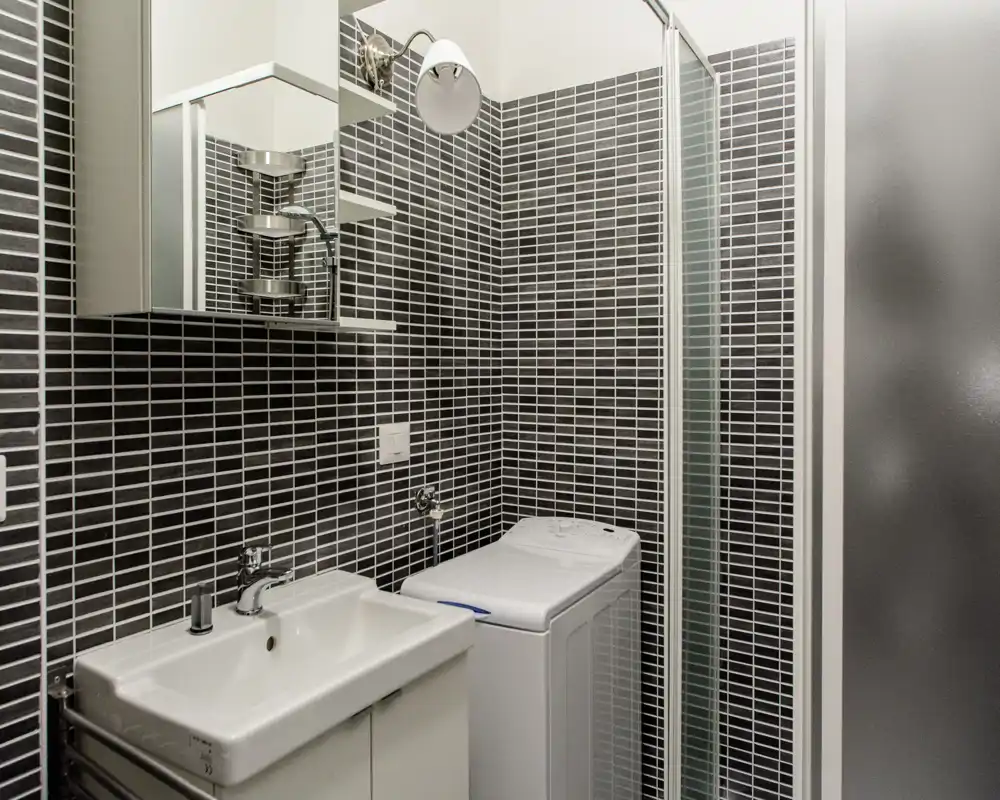 Bagno moderno e compatto con lavabo bianco, mobile specchiato, lavatrice a carica dall'alto e porta doccia in vetro su piastrelle mosaico scure.